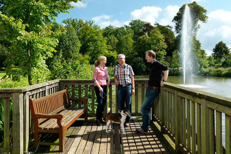 Zu sehen ist der Steg am Teich im Kaiser-Wilhelm-Hain, auf dem sich drei Menschen unterhalten