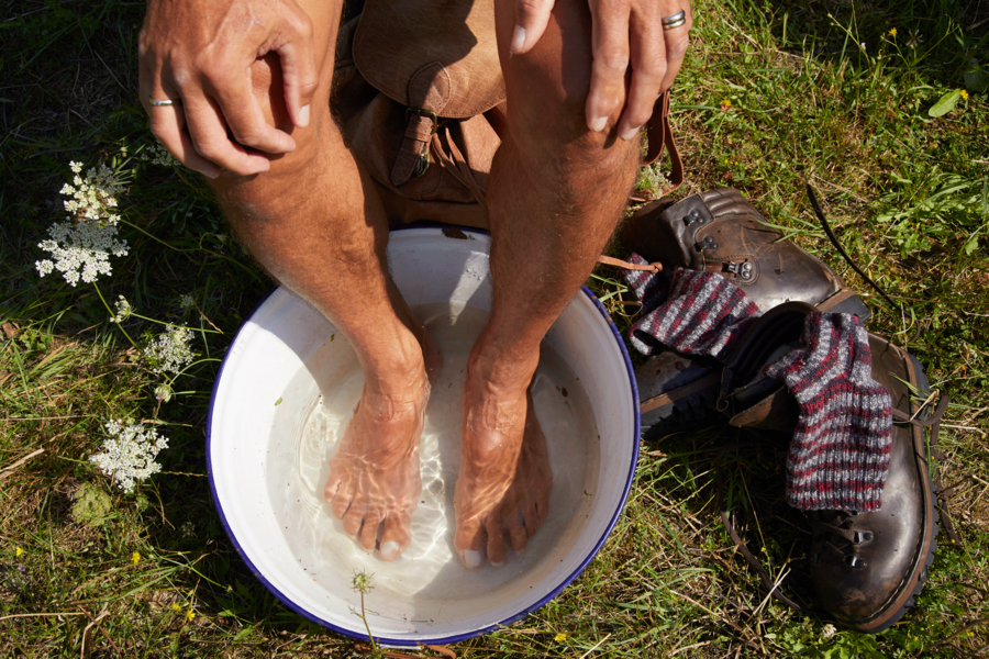 Wanderer hält nackte Füße in ein Gefäß mit Wasser, daneben liegen Wanderschuhe